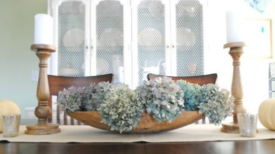 Dřevěná mísa jako dekorace pokoje plná květů hortenzie uprostřed jídelního stolu, dvě bílé svíčky na obou stranách